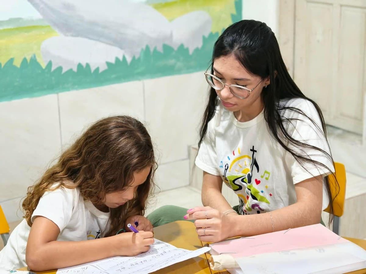 Menina escrevendo notas musicais em uma partitura ao lado de uma professora do Nosso Cantinho